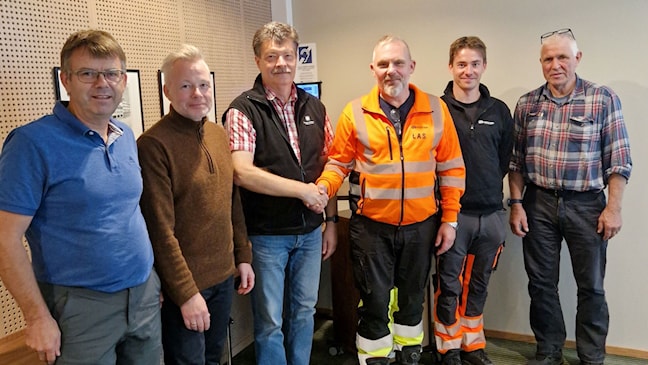 Seks menn står samlet i et rom. To tar hverandre i hånden.