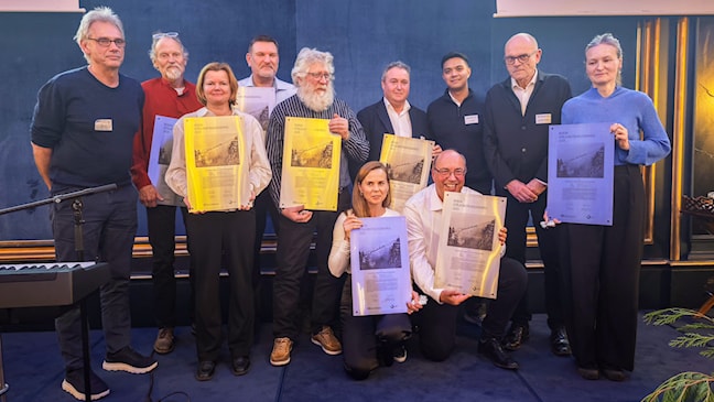11 personer, menn og kvinner, står på en scene. De holder i plaketter.