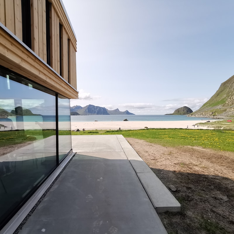 Utsnitt av trebygg med utsikt til hav, strand og fjell i horisonten.