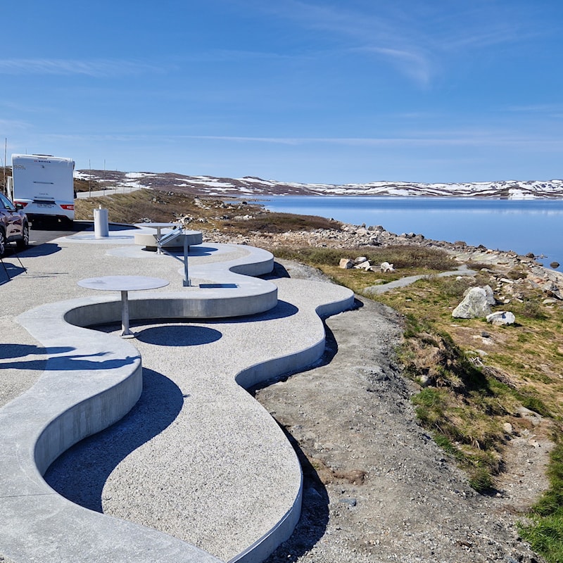 Bilde av en betongkonstruksjon som utgjør rasteområdet med bord benker. g personer som tar en pause. 